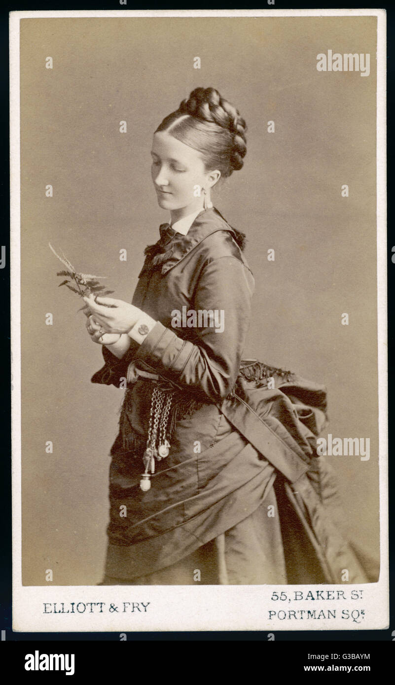 Polonaise with high bodice &amp; collar that descends to a  point behind. Hair plaited &amp;  coiled up high at the back of  the head. A chatelaine dangles  from the waist.     Date: early 1870s Stock Photo