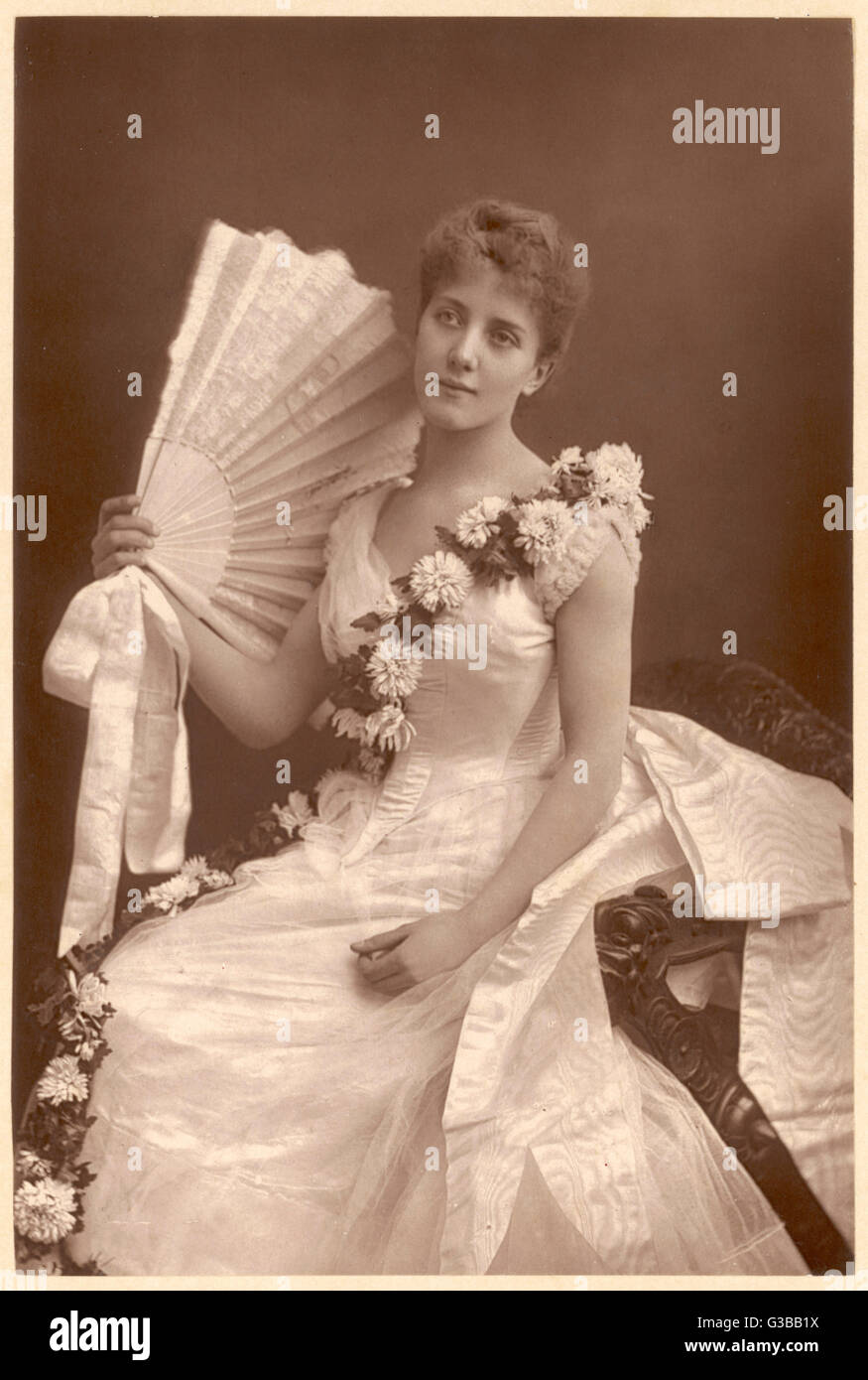 Pale sleeveless gown: pointed  waist, watered silk drapery at  the back, sash of  chrysanthemums worn across the  bodice. No jewellery but a  large lace fan with ribbon bow     Date: 1890 Stock Photo