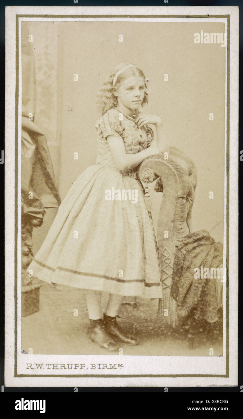 Girl wears a dress in a tiny  checked print with puffed  sleeves, round neckline,  buttoned bodice, knee-length  full skirt with narrow ribbon  trim at the hem &amp; ankle boots     Date: 1870s Stock Photo