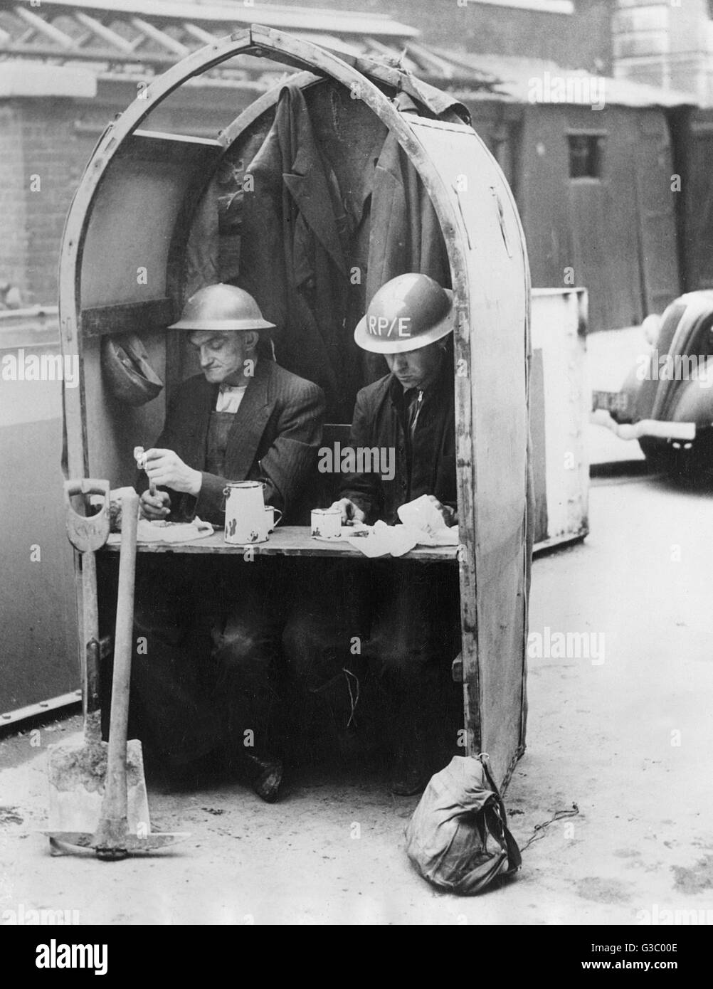 Keep Calm and Carry On. A quite fantastic photograph showing two workmen engaged in repairing air raid damage in the London area continue with their 'private' luncheon although the sirens have sounded another 'alert' - October, 1940.     Date: 1940 Stock Photo