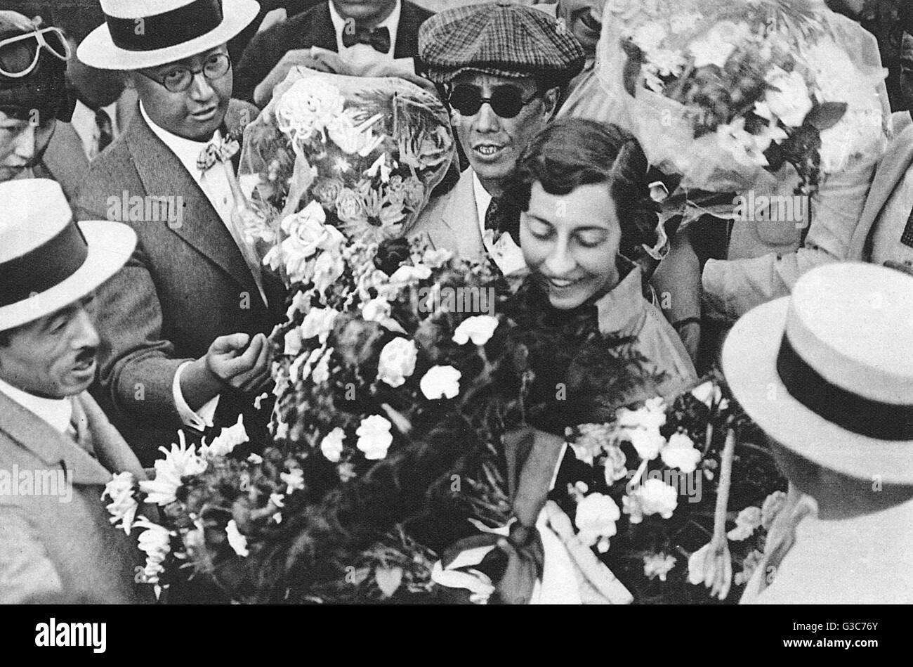 Miss Amy Johnson, the pilot, with many bouquets of flowers. She is surrounded by men, many of which wearing straw boater hats.      Date: 1932 Stock Photo