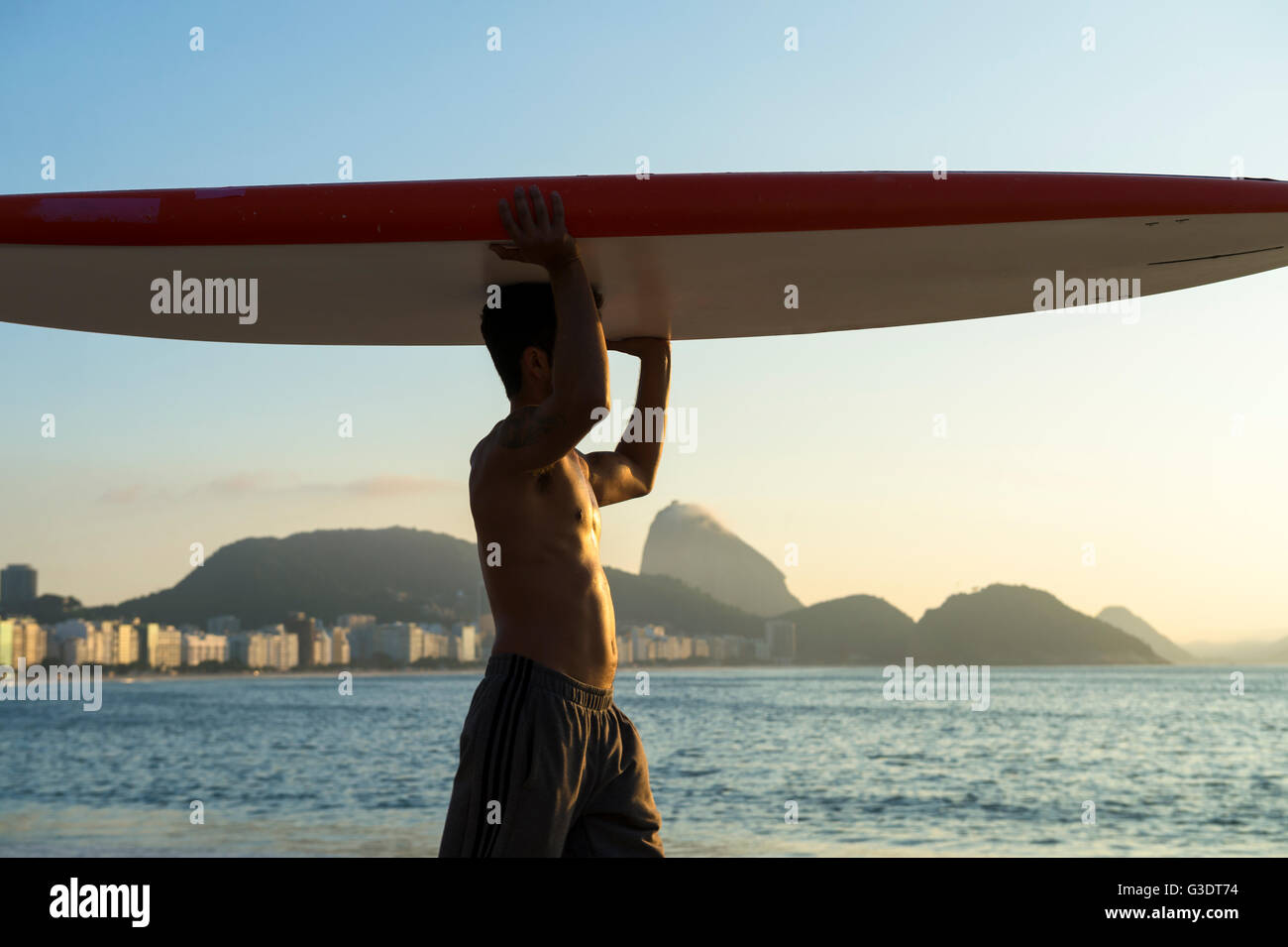RIO DE JANEIRO - APRIL 5, 2016: A young Brazilian man on Copacabana Beach carries a surfboard in front of Sugarloaf Mountain. Stock Photo