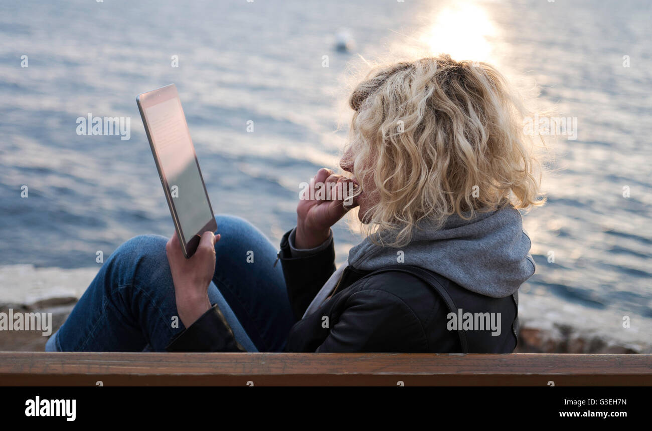 Blonde girl with curly hair is seating on the bench and absorbedly watching something in tablet Stock Photo