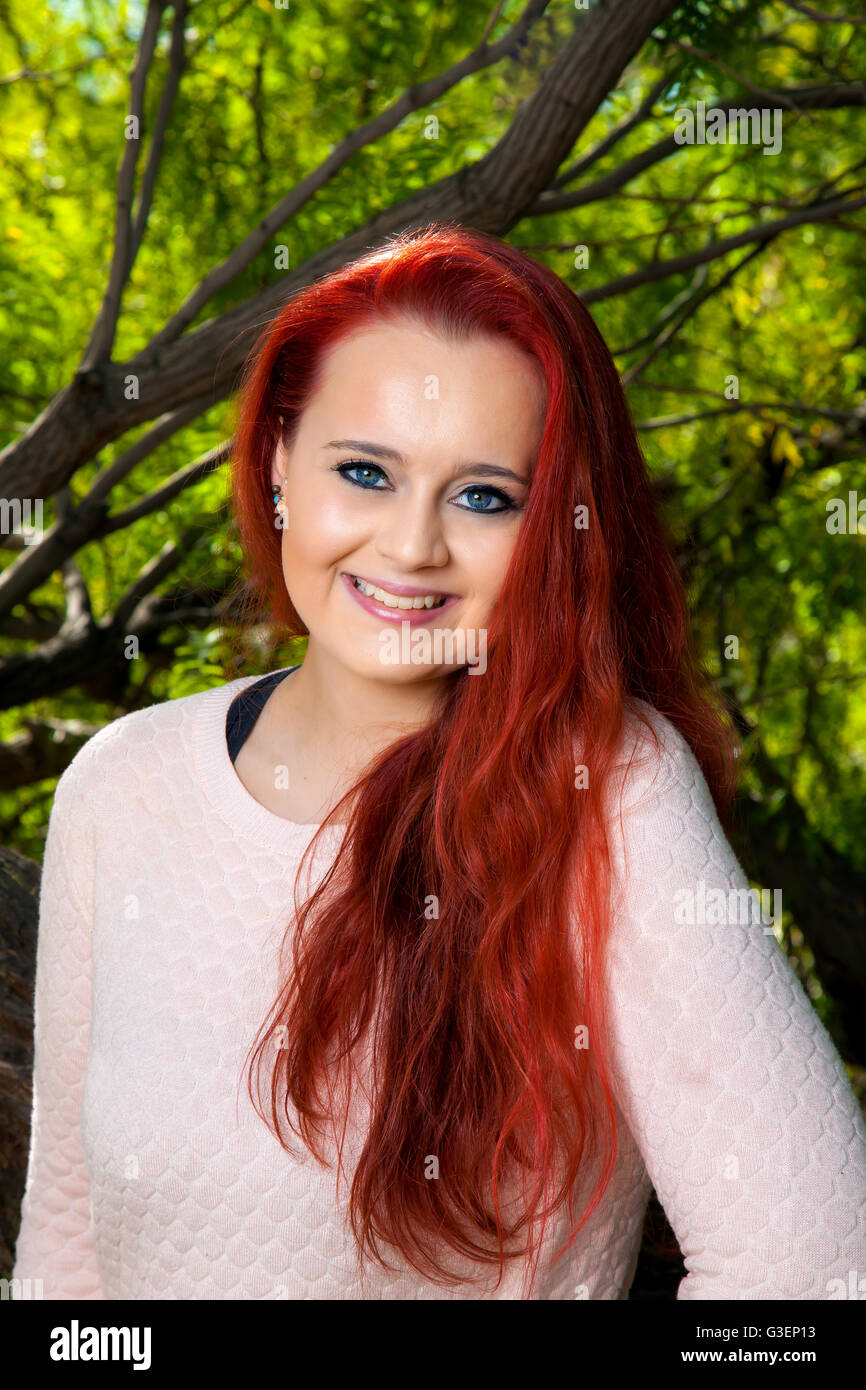 A beautiful, teenage girl with dyed red hair poses for a portrait in front of trees.  She has a tilted head, lovely smile, and Stock Photo