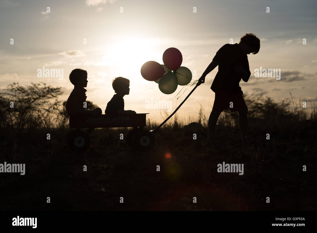 Three boys in silhouette playing with cart on hill Stock Photo