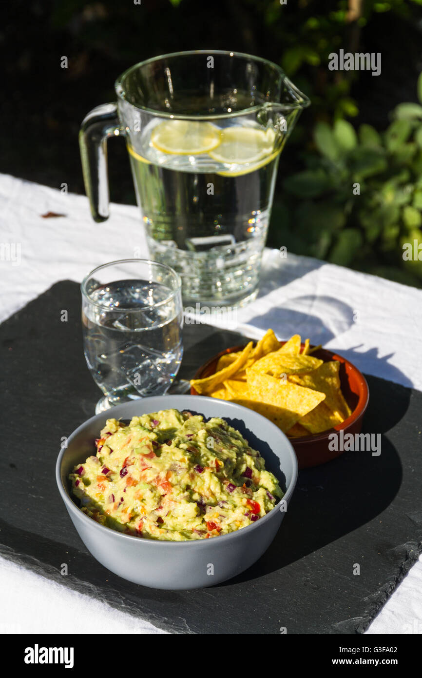 Home made guacamole in ceramic bowl and tortilla chips and soured cream on the side with jug and glass of iced water. On slate m Stock Photo