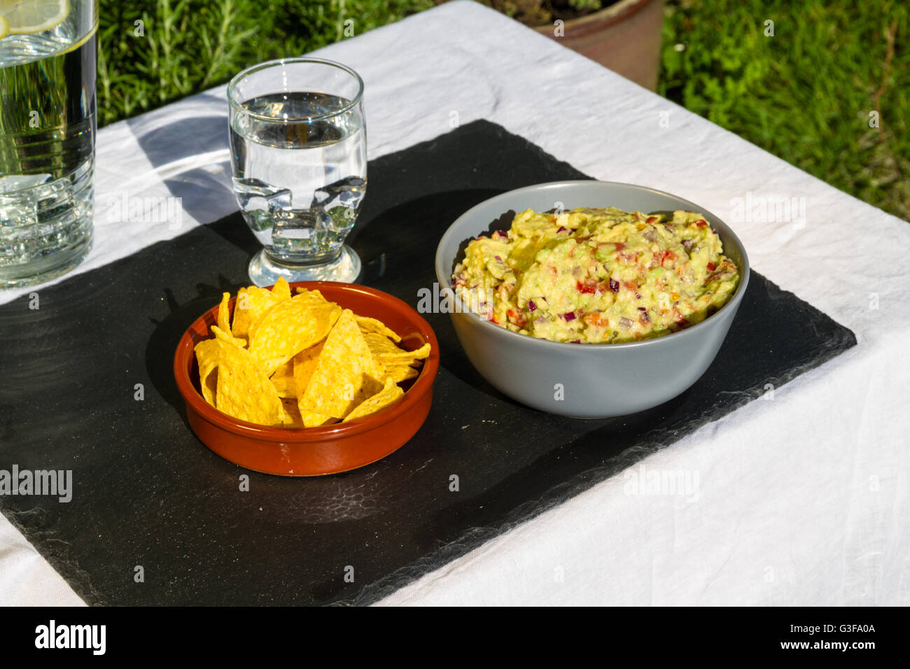 Home made guacamole in ceramic bowl and tortilla chips and soured cream on the side with jug and glass of iced water. On slate m Stock Photo