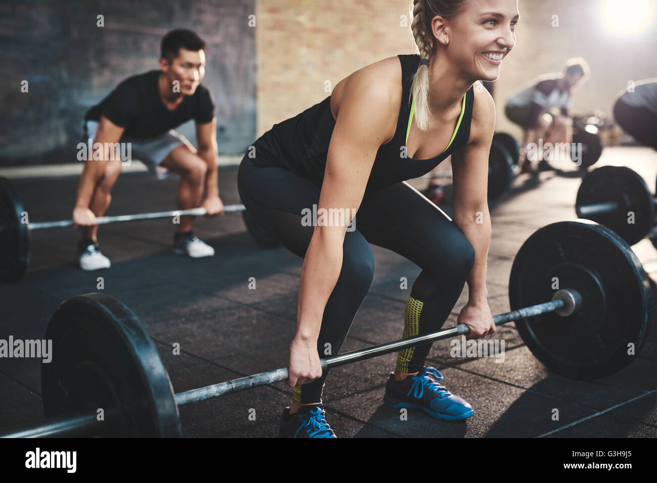 Cheerful muscular young woman with ponytail and black tights performing dead lift barbell exercises with other students Stock Photo