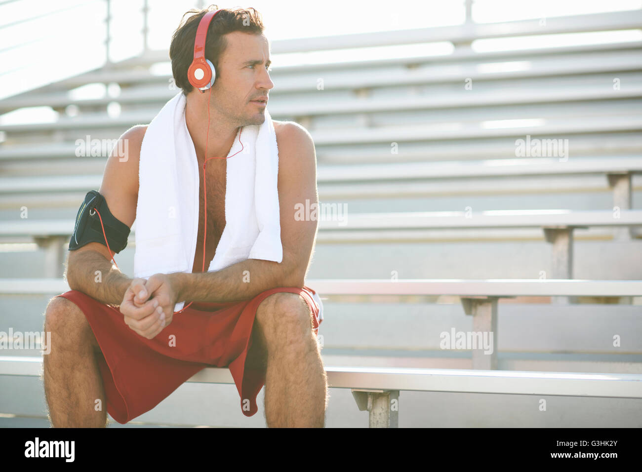 Mid adult man sitting on bench, taking a break from exercise, wearing headphones, towel around neck Stock Photo