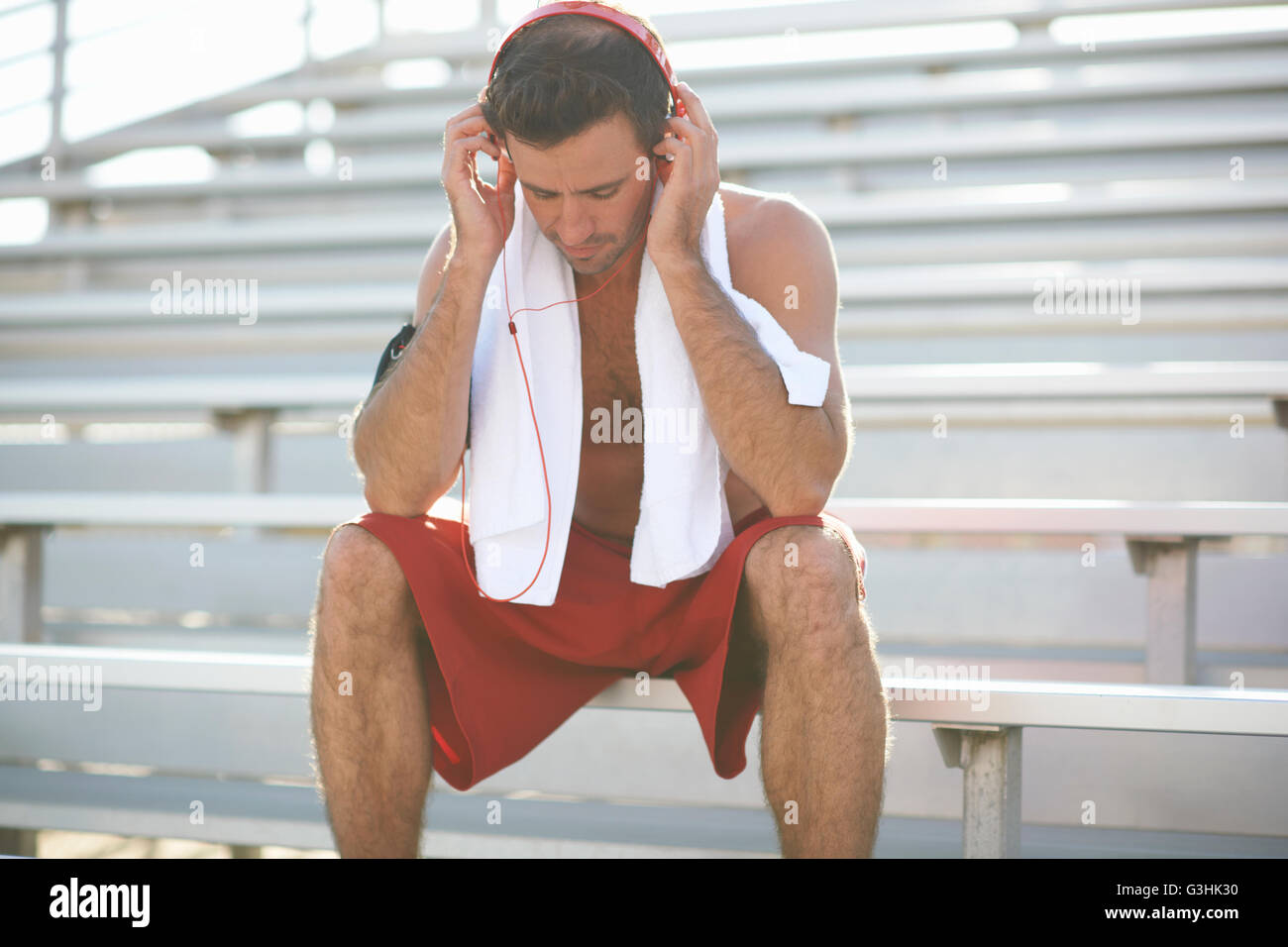 Mid adult man sitting on bench, taking a break from exercise, wearing headphones, towel around neck Stock Photo