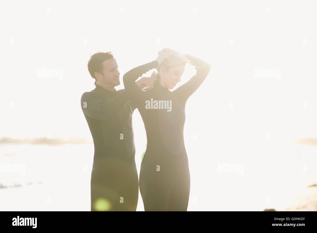 Surfing couple zipping up wetsuits on sunlit Venice Beach, California, USA Stock Photo