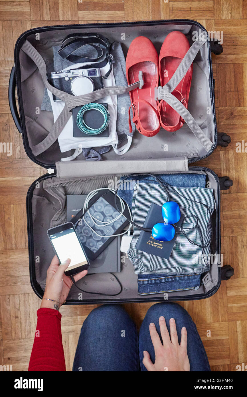 Woman packing suitcase, holding smartphone, overhead view Stock Photo