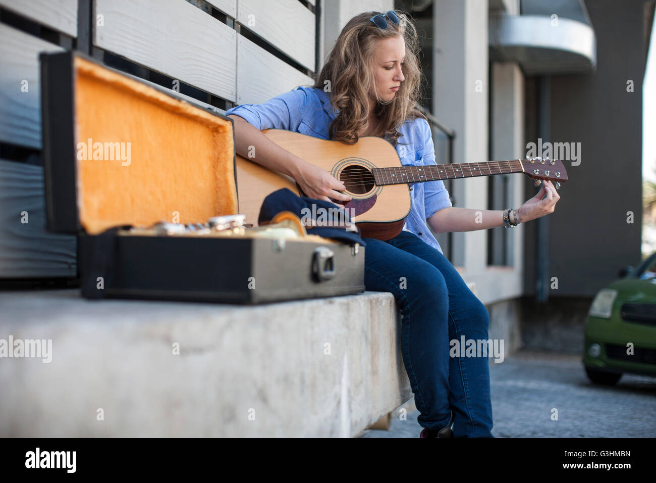 Female street musician sitting on ledge, tuning guitar Stock Photo