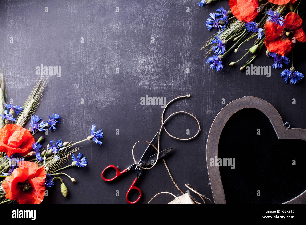 Red poppies with blue cornflowers and rye on the corners of old blackboard with scissors and wooden heart Stock Photo