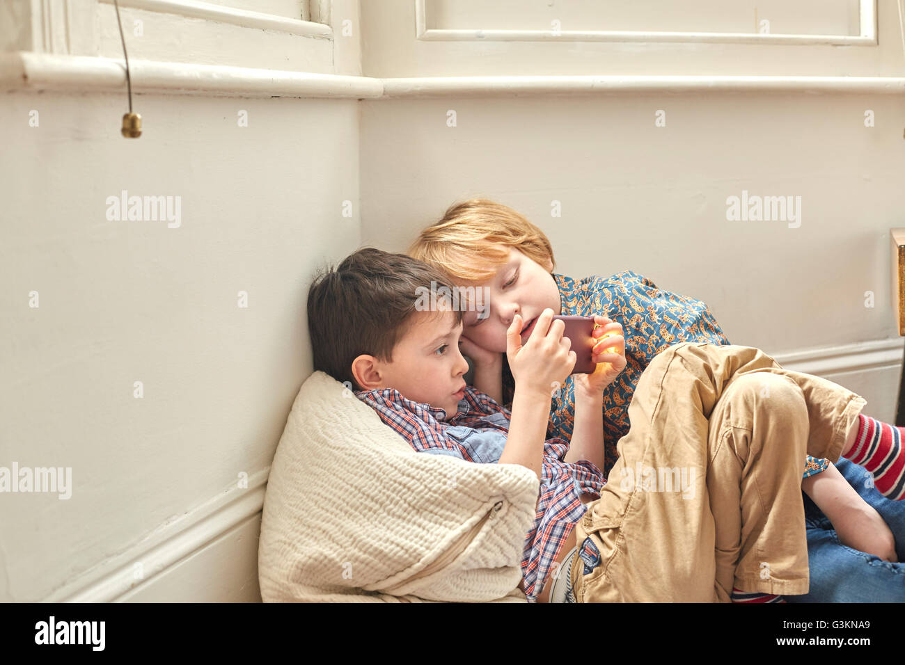 Two young boys leaning, sitting indoors, against wall, looking at smartphone Stock Photo