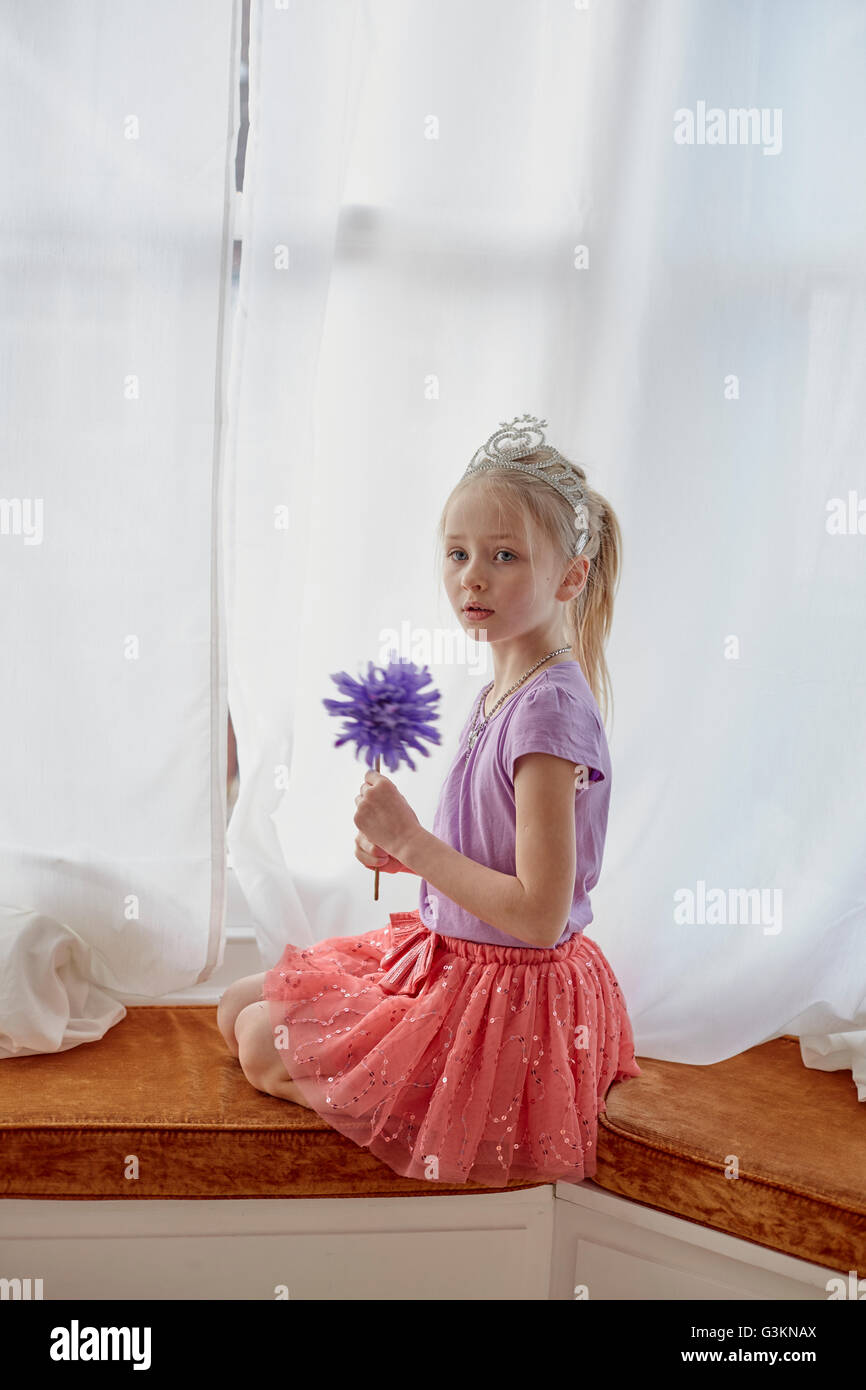 Portrait of young girl sitting by widow, wearing tiara, holding wand Stock Photo