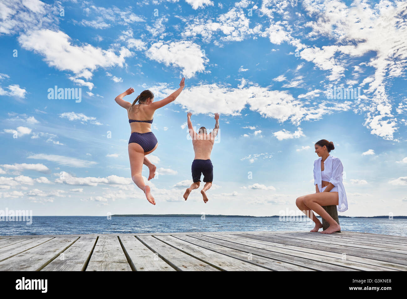 Young woman sitting on post on wooden pier, watching friends jump into sea, rear view Stock Photo