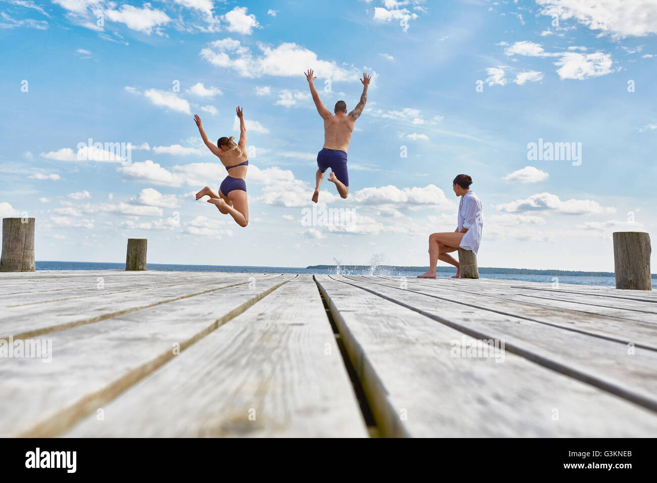 Young woman sitting on post on wooden pier, watching friends as they jump into sea Stock Photo