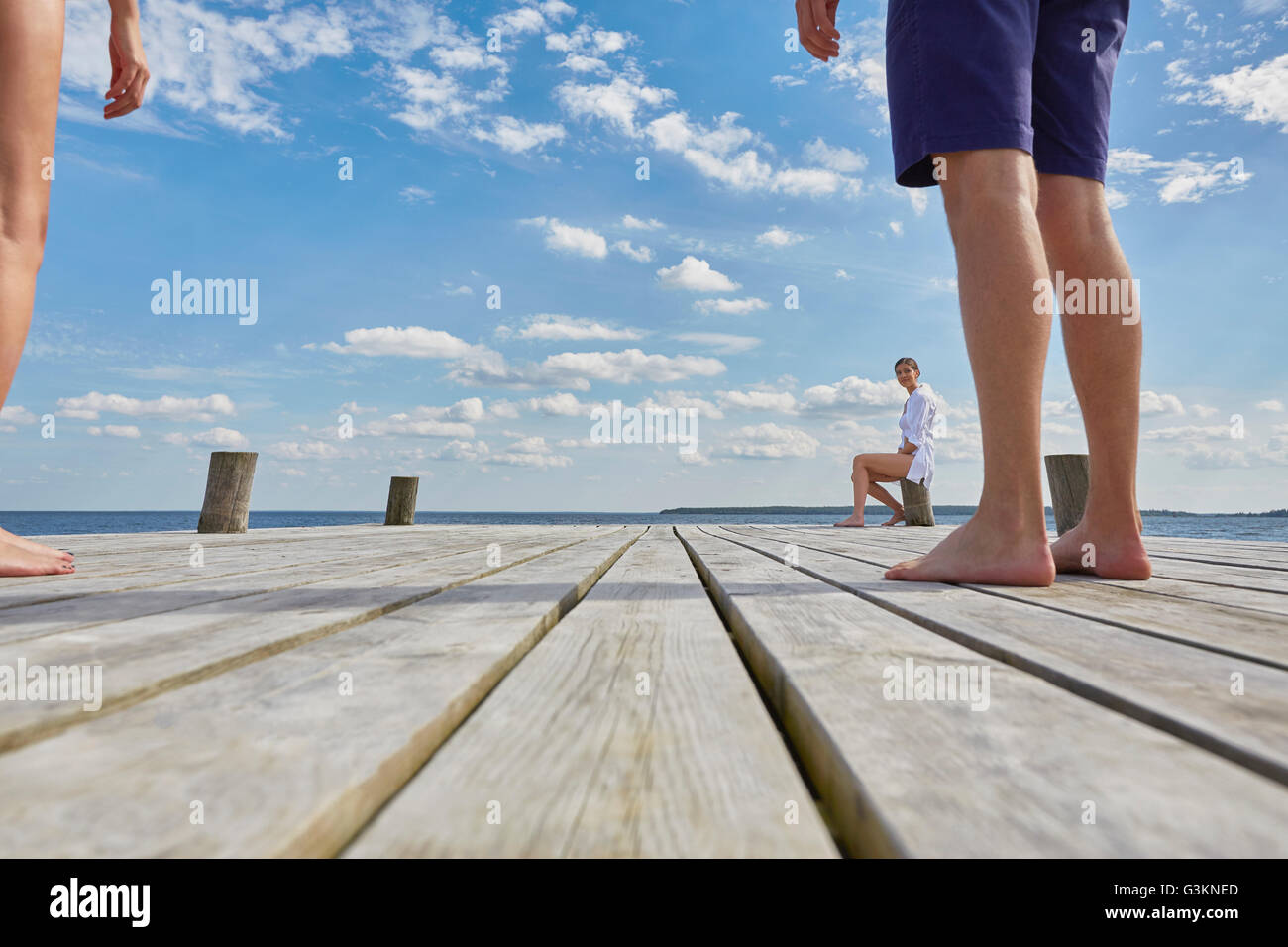 Young woman sitting on post on wooden pier, looking at friends standing further away Stock Photo