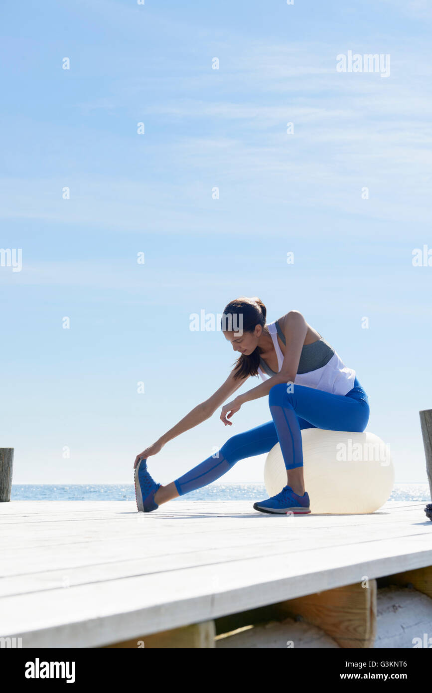 Women wearing sports clothes sitting on pier on exercise ball stretching foot Stock Photo