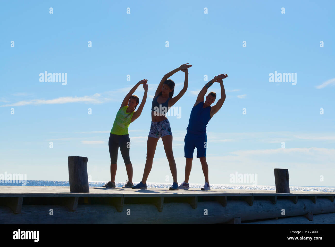Friends on pier wearing exercise clothes arms raised, bending over sideways stretching Stock Photo