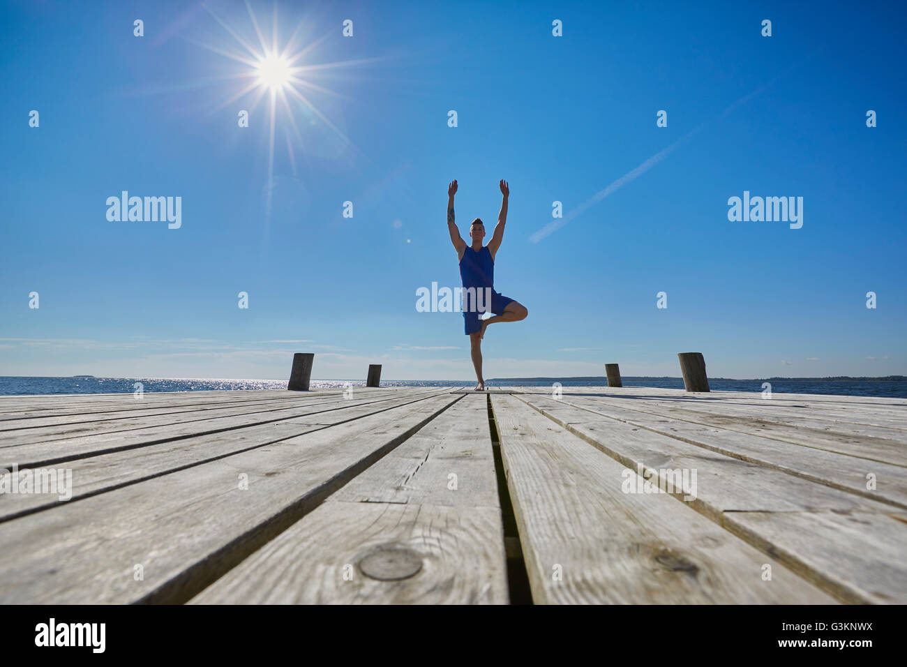 Mid distant view of man on pier, standing on one leg arms raised Stock Photo