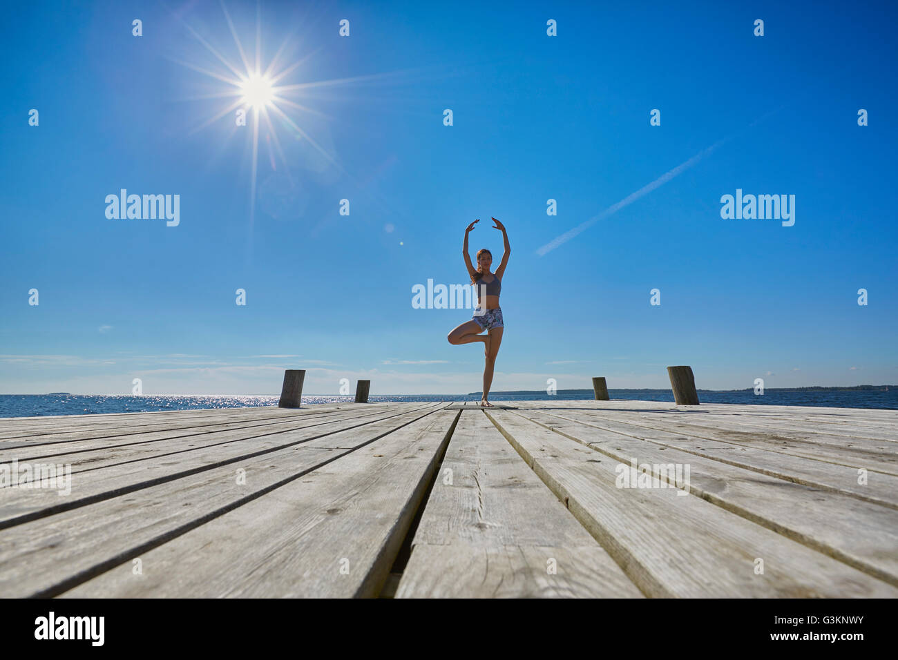 Mid distant view of woman on pier, standing on one leg arms raised Stock Photo
