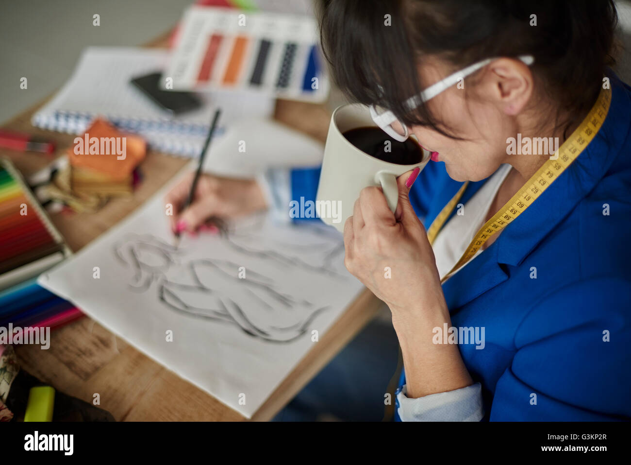 Over shoulder view of woman in design studio drinking coffee, sketching fashion design Stock Photo
