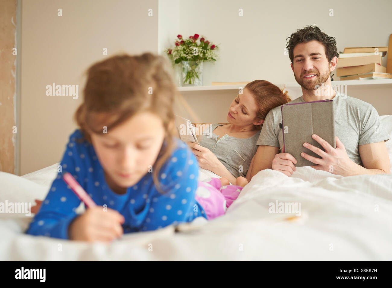Mid adult couple having lie in whilst daughter draws on bed Stock Photo