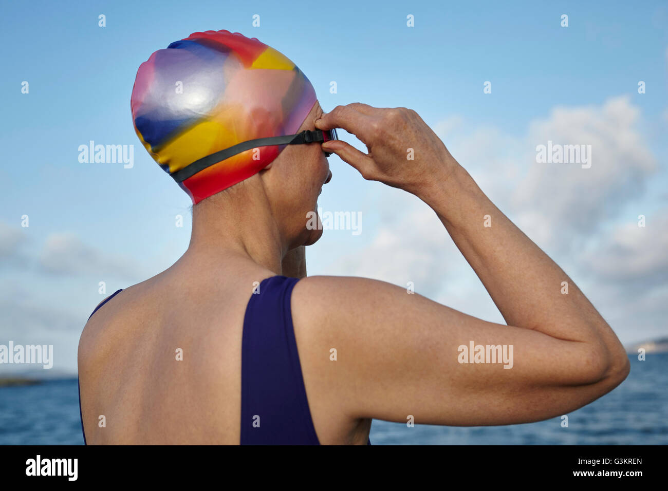 Mature woman wearing swimming costume and swimming hat, standing beside the sea, adjusting swimming goggles, rear view Stock Photo