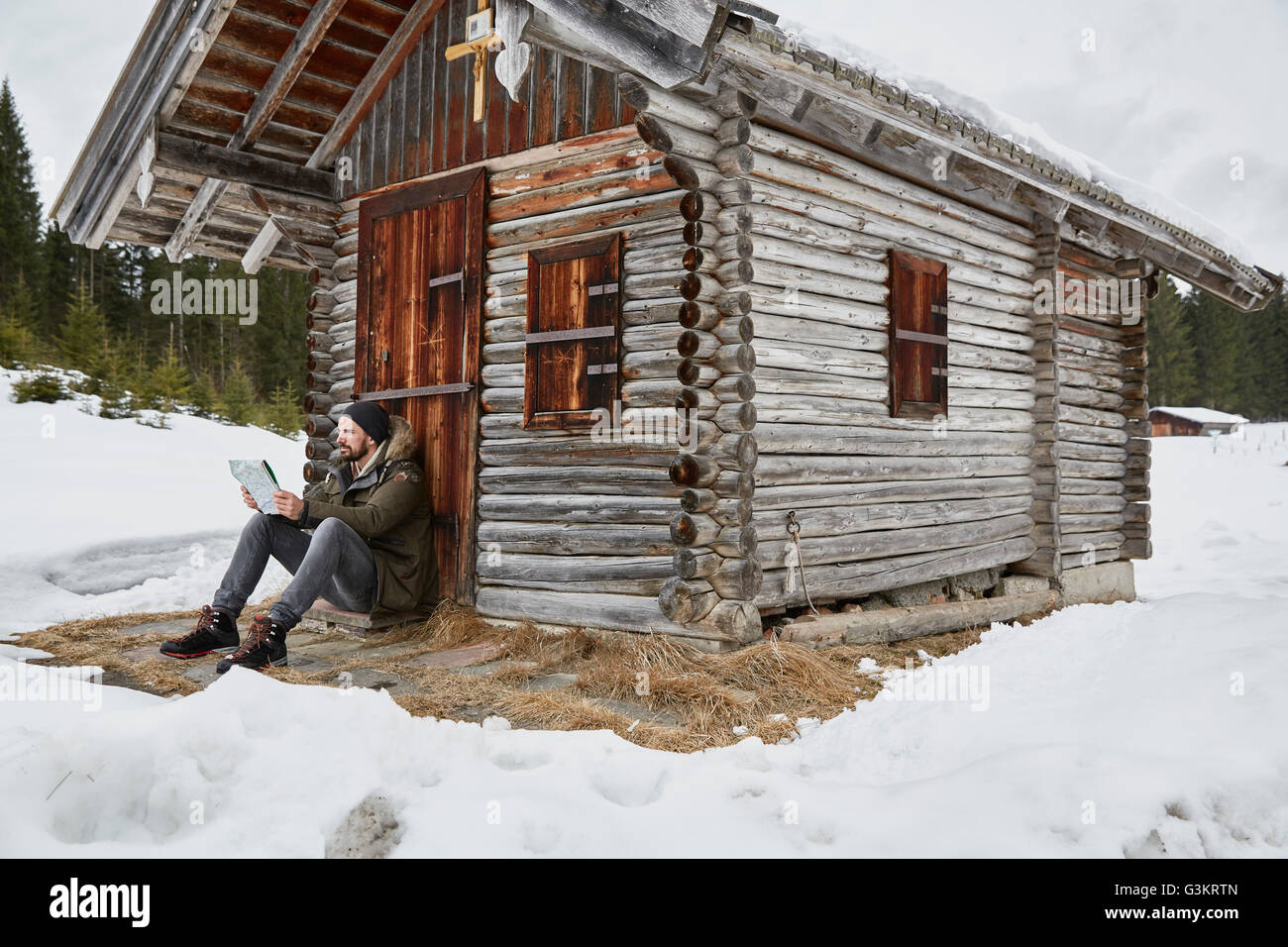 Young man reading map sitting outside log cabin in winter, Elmau, Bavaria, Germany Stock Photo