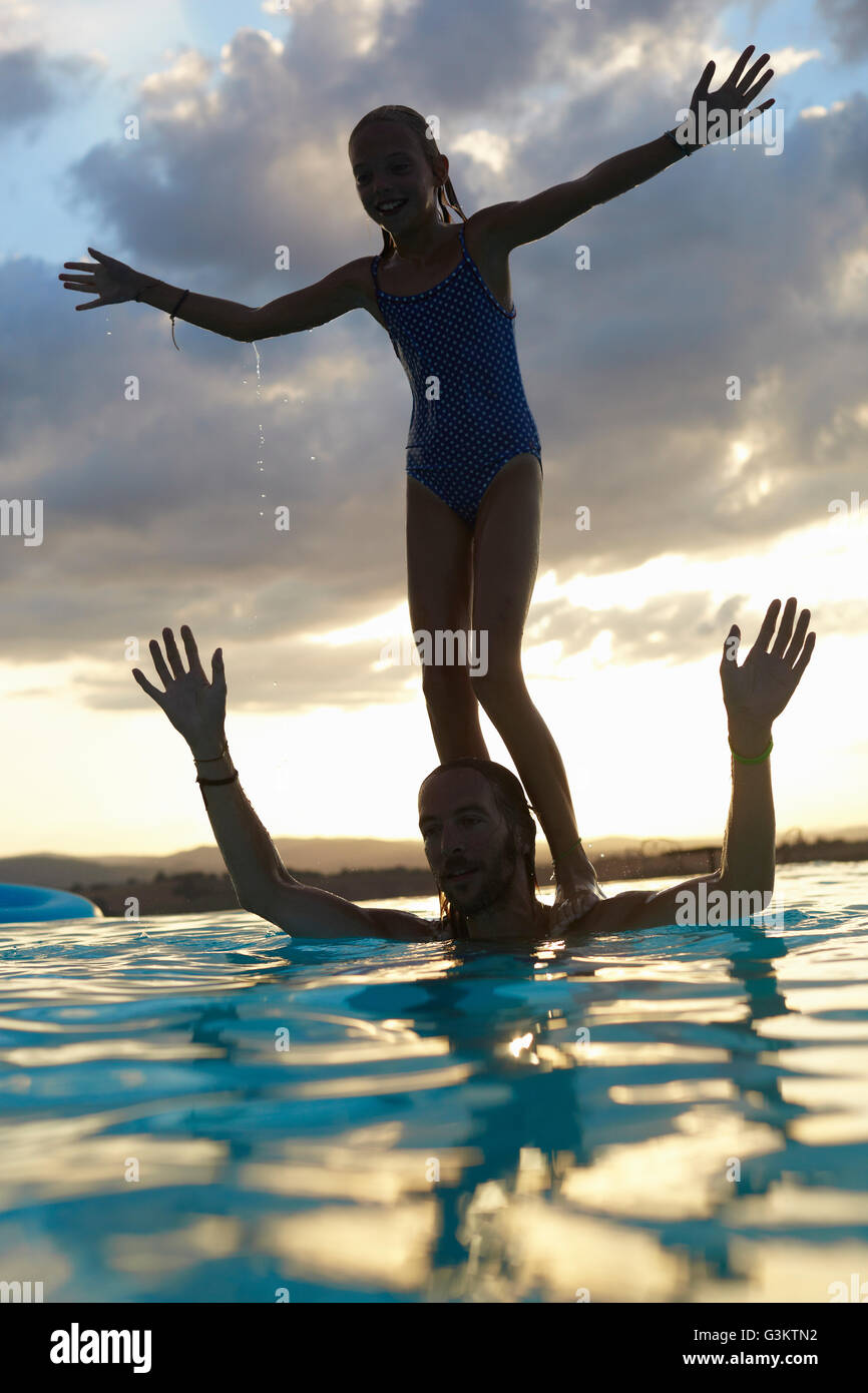 Silhouetted girl balancing on fathers shoulders in swimming pool, Buonconvento, Tuscany, Italy Stock Photo
