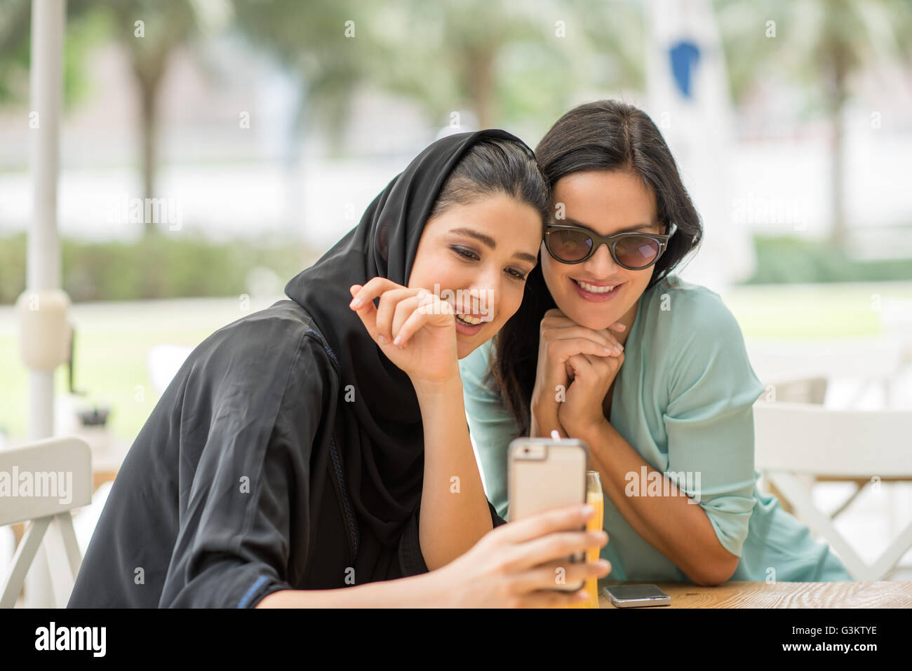 Young middle eastern woman wearing traditional clothing reading smartphone text with female friend at cafe, Dubai, United Arab Emirates Stock Photo