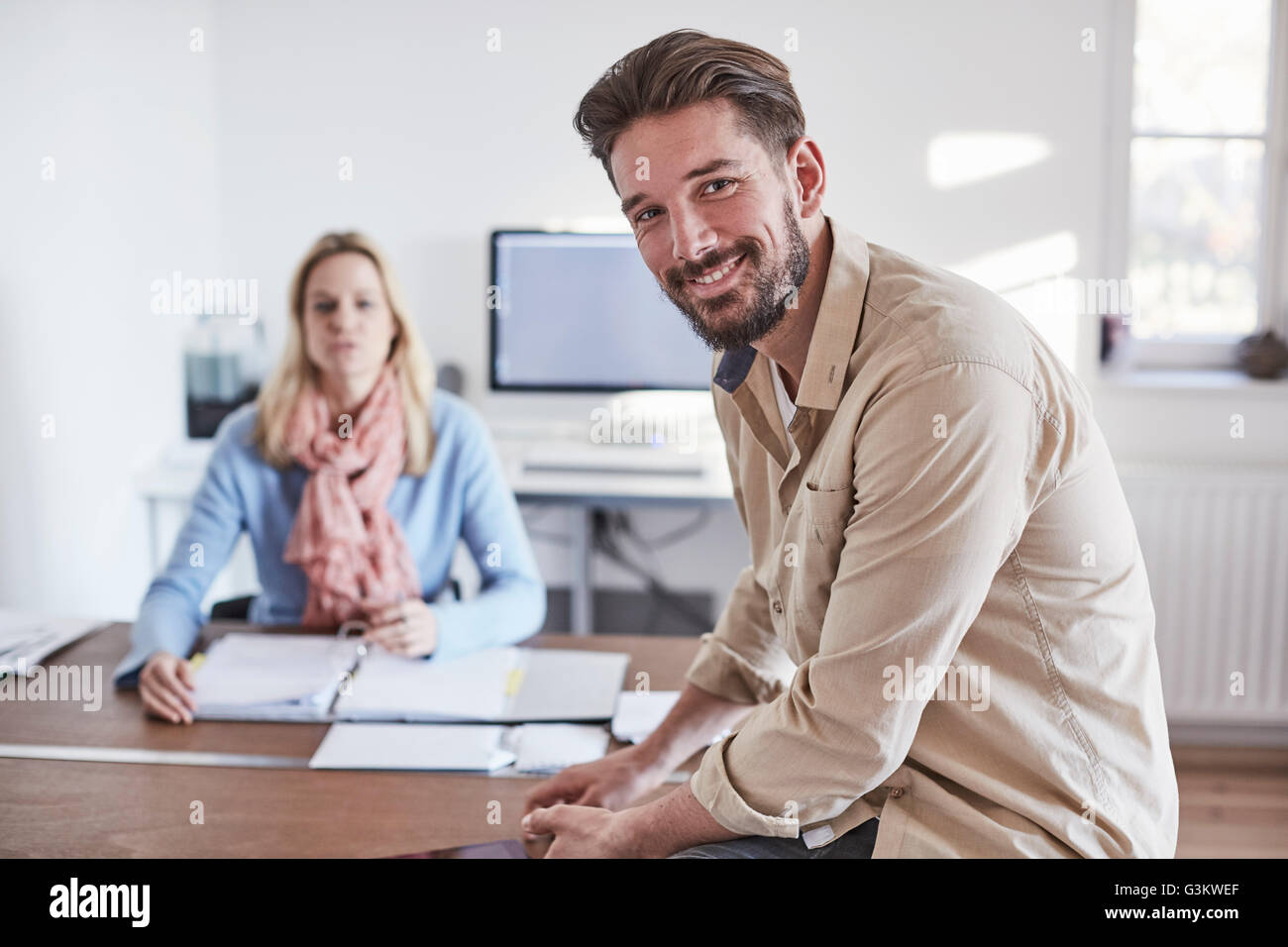 Man sitting on desk in office looking at camera smiling Stock Photo