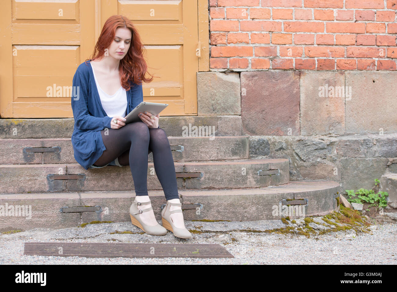 Young woman sitting on stairs in front of building holding a tablet pc ...