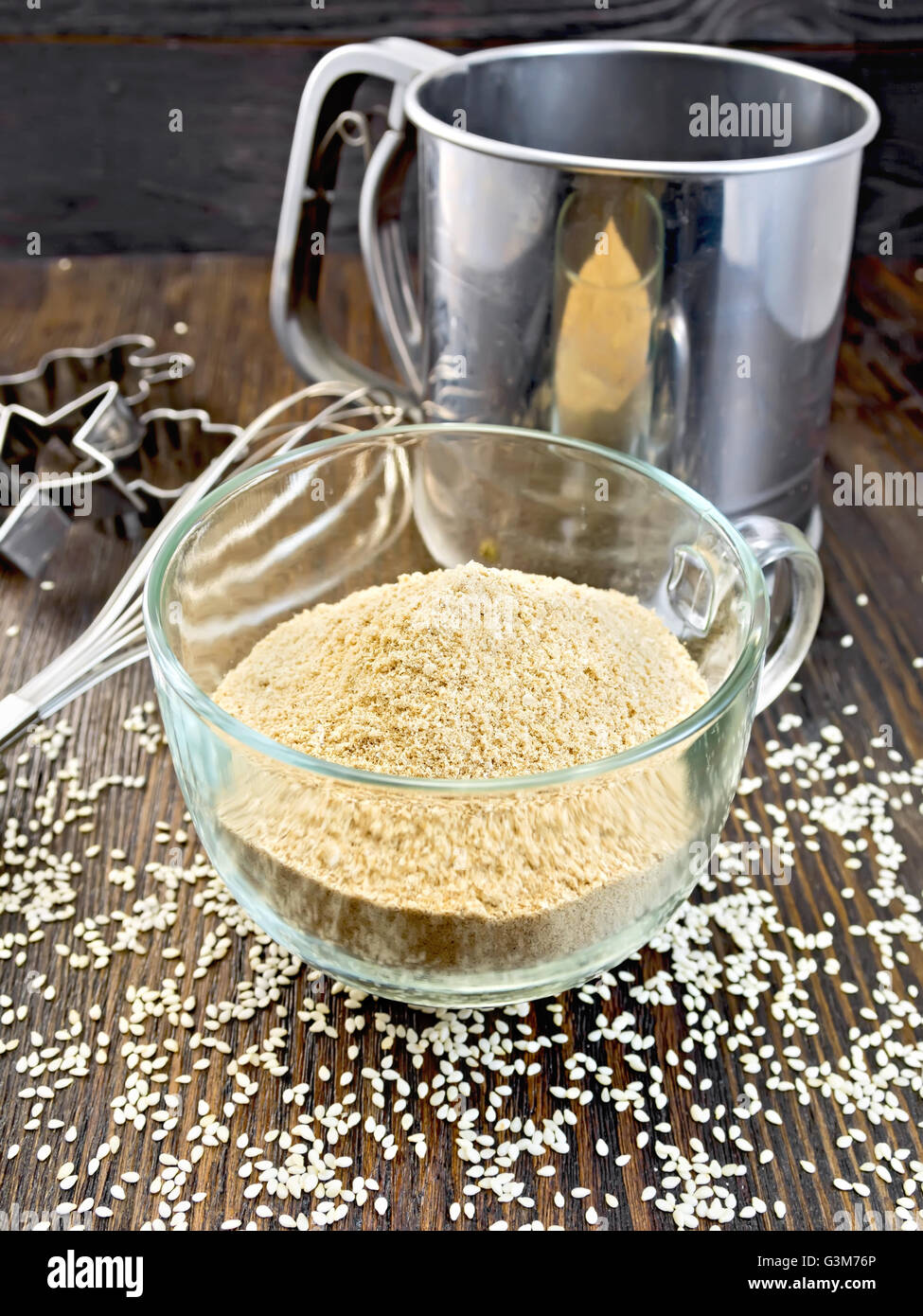 Sesame flour in a glass cup with strainer and mixer, sesame seeds on a wooden boards background Stock Photo