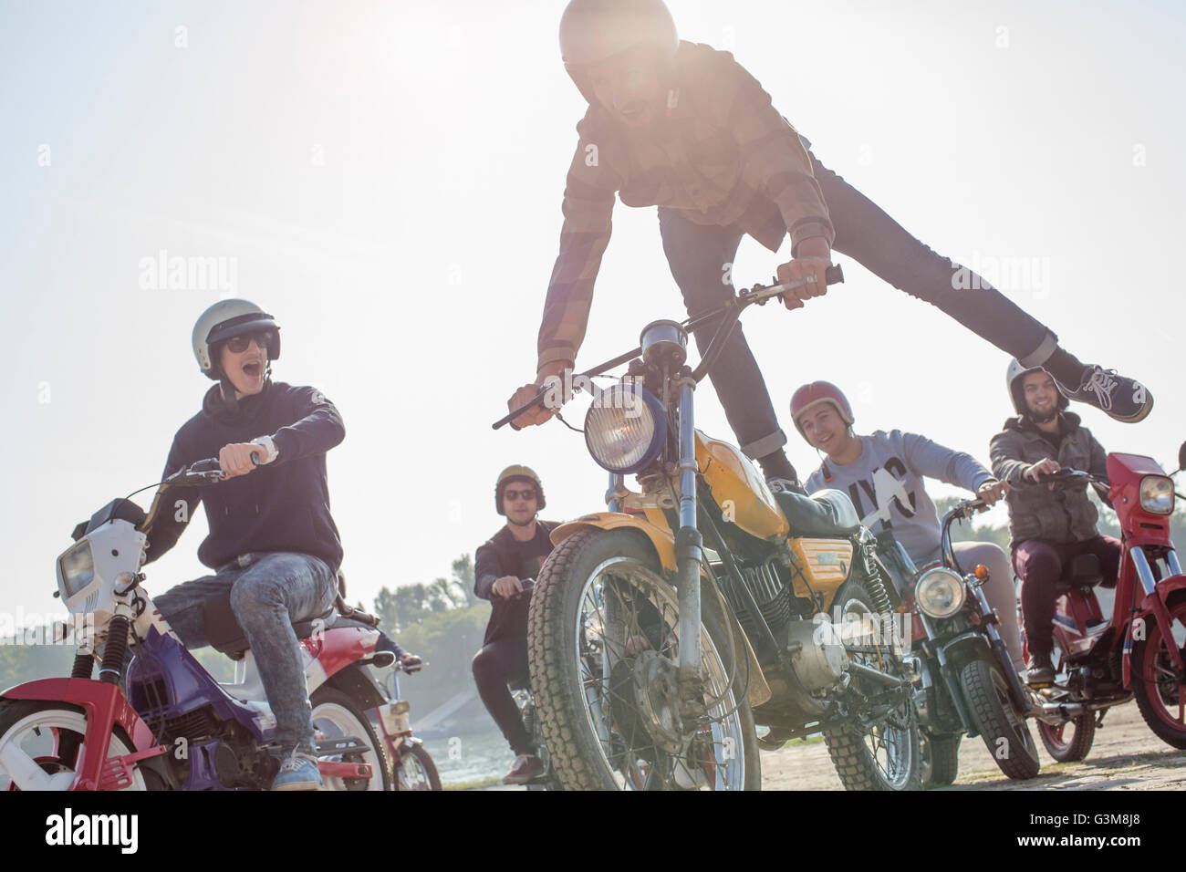 Group of friends riding mopeds along road, man in mid air, doing stunt Stock Photo