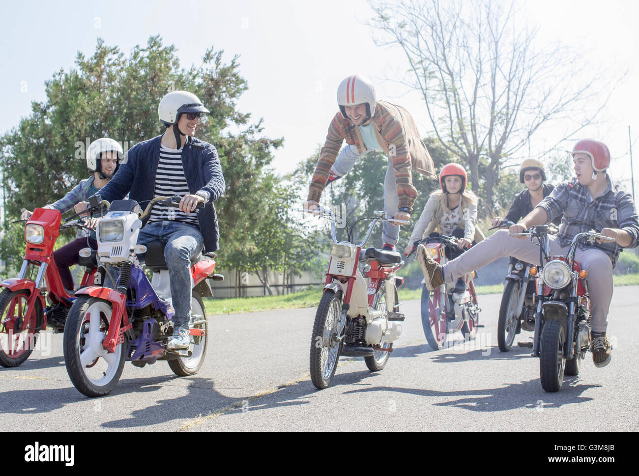 Group of friends riding mopeds along road, man in mid air, doing stunt Stock Photo