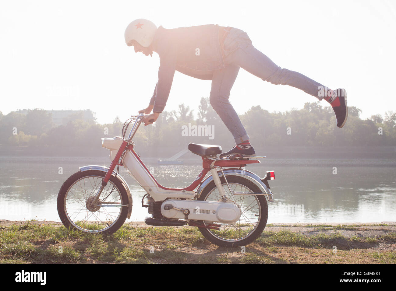 Mid adult riding moped, doing stunt, beside lake Stock Photo