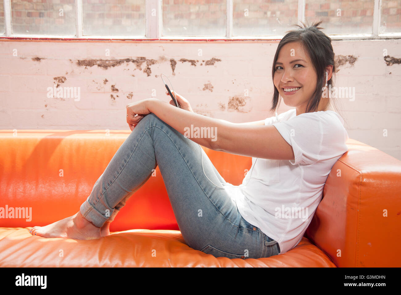 asian woman listening to music on her ipad sitting on a orange sofa Stock Photo
