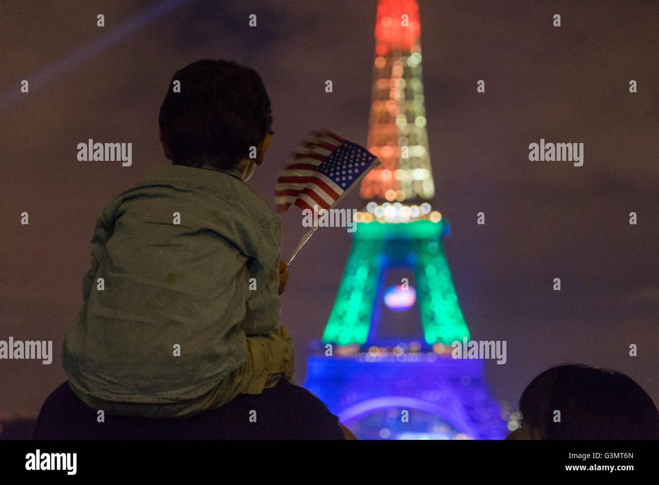 Paris, France. 13, June, 2016. A young child sitting on the shoulders of his father waves an American flag while watching the Eiffel Tower lit with rainbow colors to show support to the victims of the Orlando shooting. Credit:  David Bertho / Alamy Live News Stock Photo