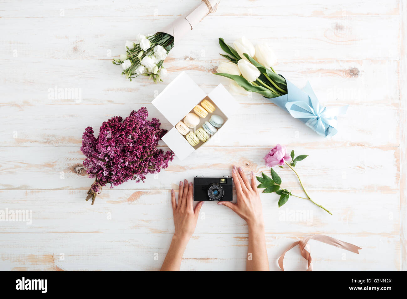 Hands of young woman holding photo camera on wooden table with french macaroons and flowers Stock Photo