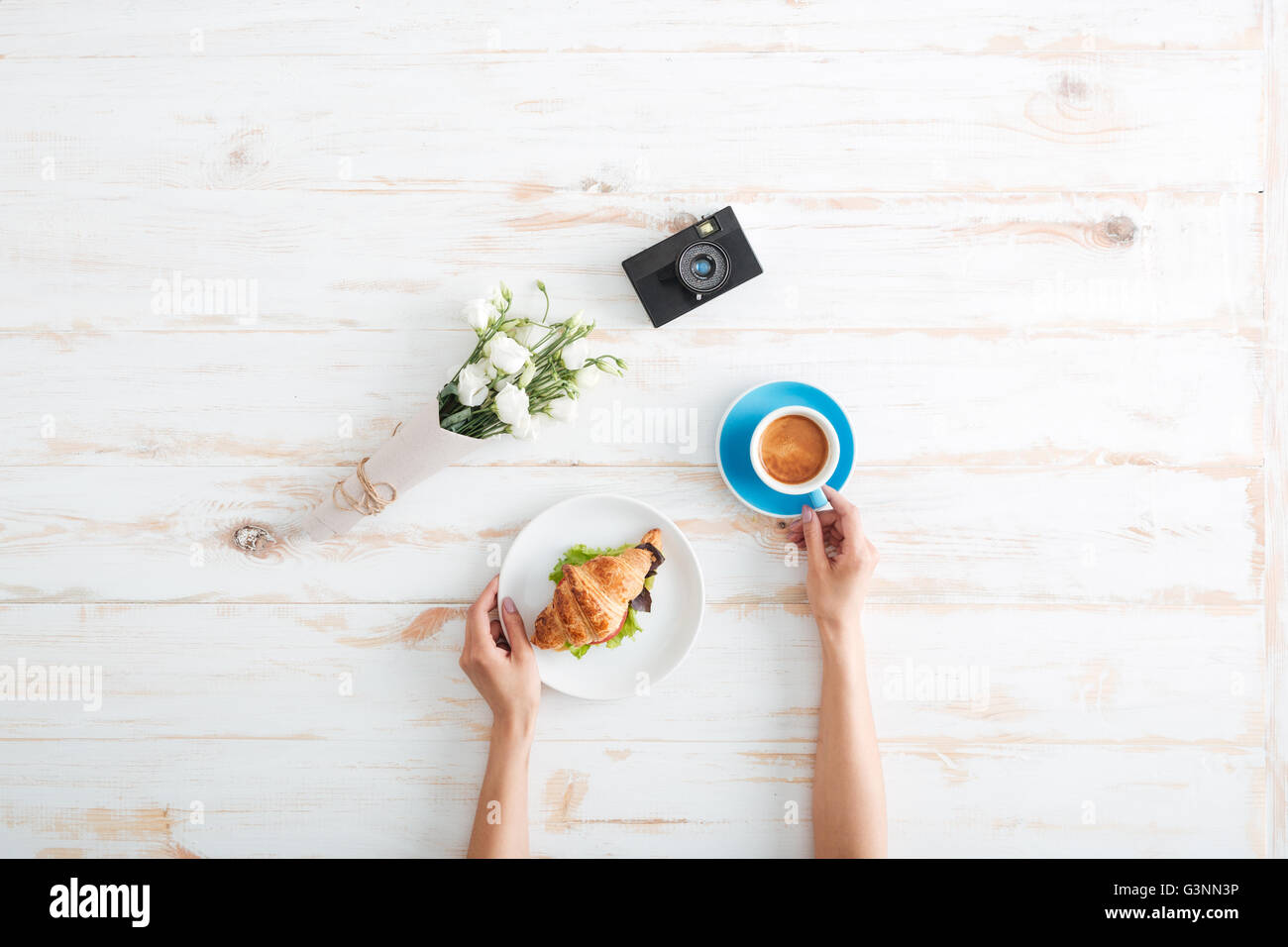 Hands of young woman drinking coffee with croissant on wooden table with flower bouquet and photo camera Stock Photo