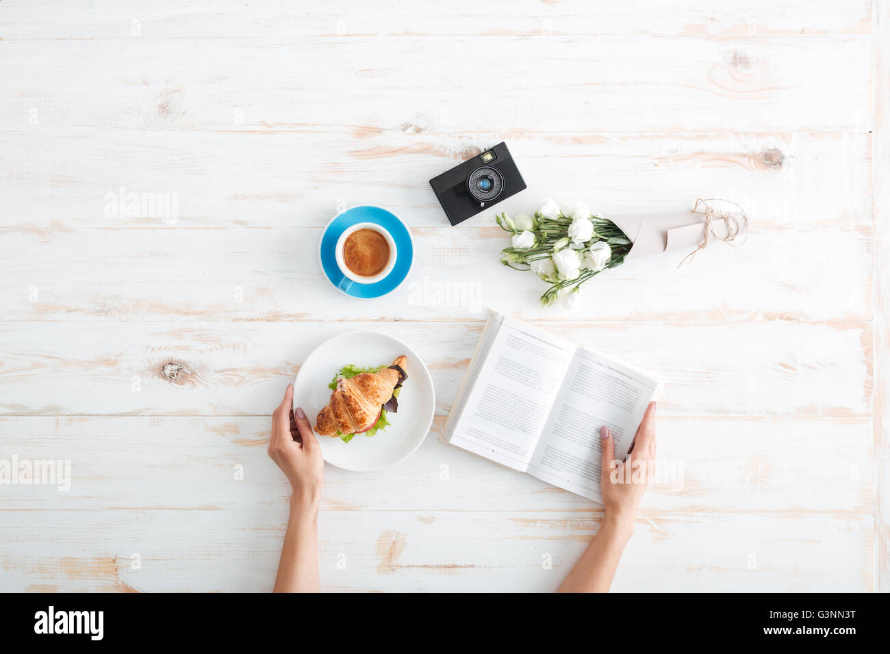 Hands of young woman eating croissant with coffee and reading book on wooden table with flower bouquet and photo camera Stock Photo
