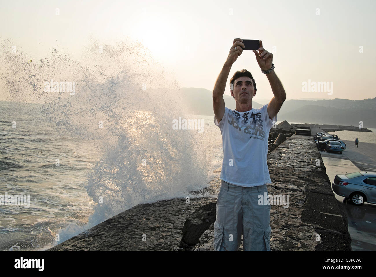 A middle aged man captures a selfie of himself in front of a wave crashing onto rocks in Amasra, Bartin, Turkey Stock Photo