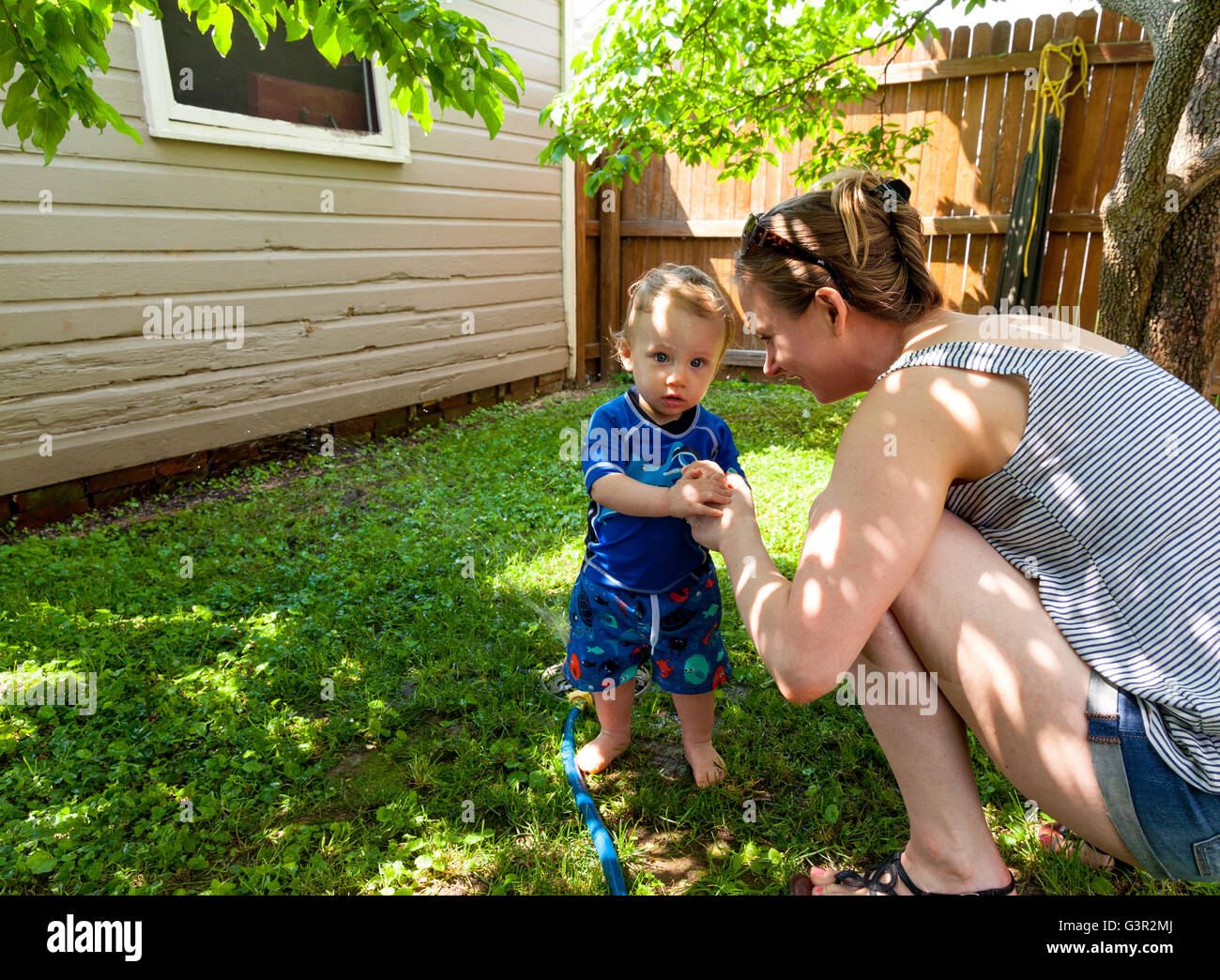 Baby Caucasian Boy and Mother playing in his backyard with a garden sprinkler Stock Photo