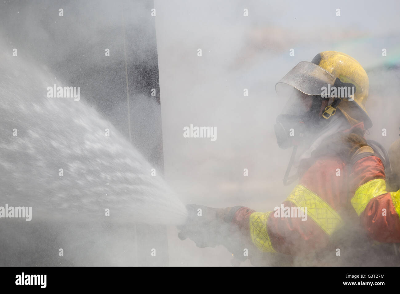 fireman in helmet and oxygen mask  spraying water to fire surround with smoke and drizzle Stock Photo