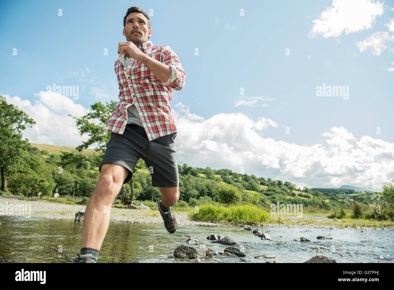 A man in a red check shirt striding across a shallow river near Bala Lake in Wales. Stock Photo