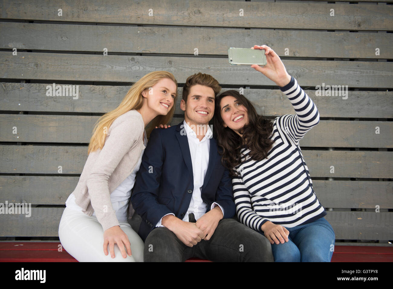Three friends sitting on a bench taking a selfie on the South Bank in London. Stock Photo