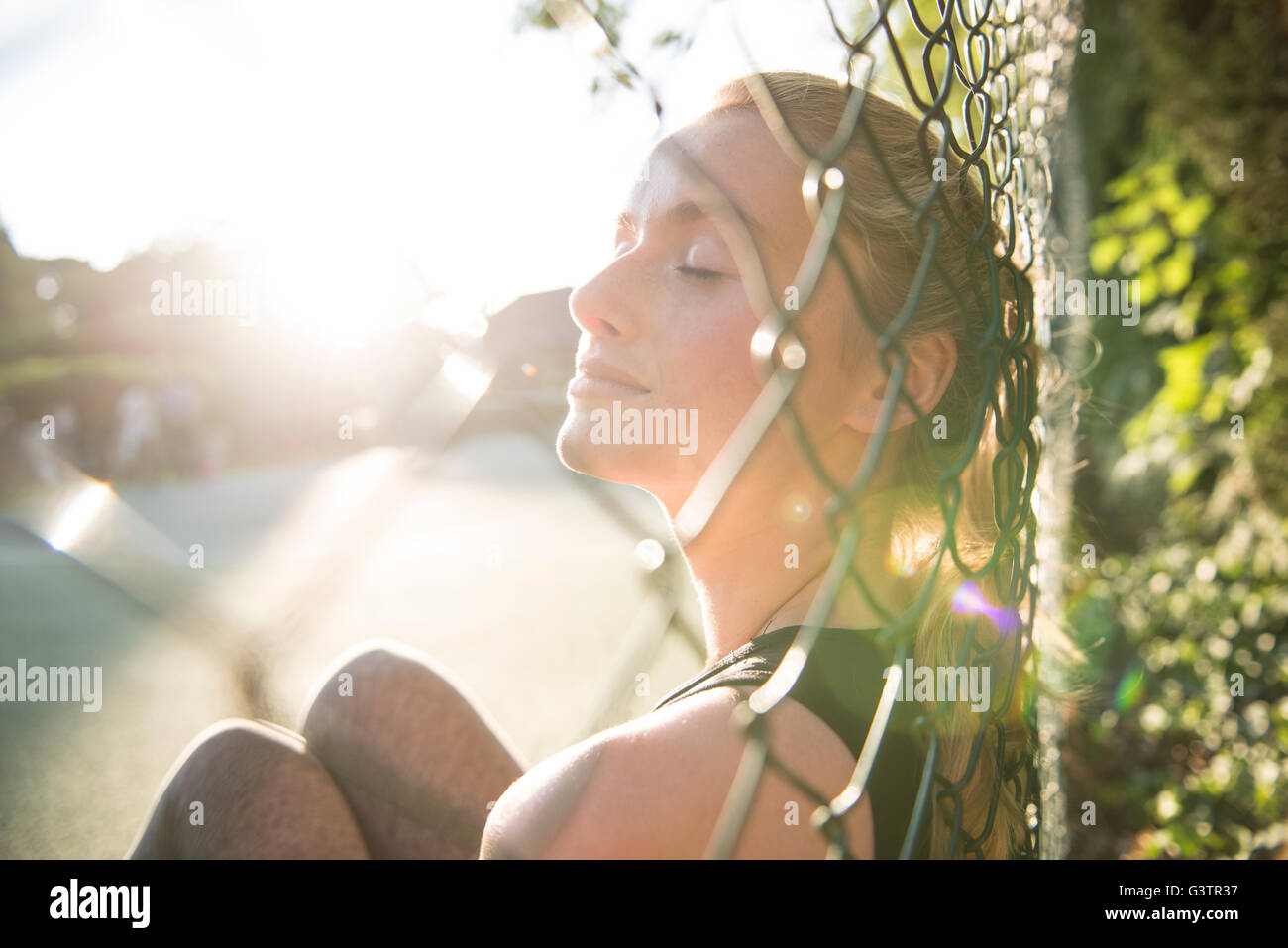 A young woman enjoying the evening sunshine sat on a tennis court. Stock Photo